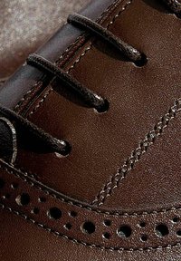 Scarpa in pelle marrone con lacci neri, caratterizzata da dettagli perforati lungo il bordo e cuciture a contrasto. Texture liscia.