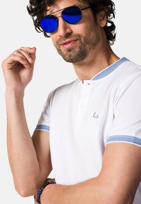 Biała koszulka polo z niebieskim akcentem i zapięciem na dwa guziki. Model nosi niebieskie okulary przeciwsłoneczne z lustrzanym filtrem i bransoletkę z koralików na nadgarstku.