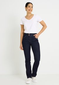 Vaqueros azul oscuro de tiro alto, con dos botones dorados y bolsillos delanteros. Combinados con una camiseta blanca lisa de cuello en V y zapatos blancos.