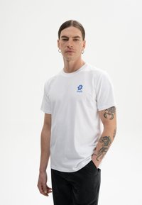 Witte katoen t-shirt met een blauw bloemenontwerp en tekst op de linkerborst. Het model heeft tatoeages en draagt zwarte broek.