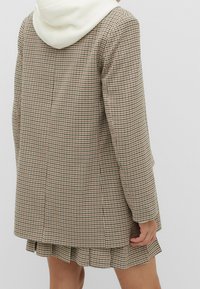 Blazer à carreaux marron et crème avec une grande capuche, présentant une coupe décontractée, une texture lisse, et une jupe plissée en dessous.