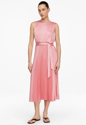Kobieta w różowej, bezrękawowej sukience midi z plisowaną spódnicą i dopasowanym paseczkiem w talii, w czarnych sandałach, stojąca na białym tle.