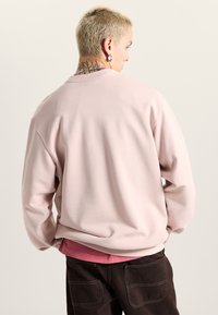 Lyserød sweatshirt i afslappet pasform, ribbet krave og lange ærmer, lagdelt over en maroon skjorte. Tekstureret stof med minimal detaljering.
