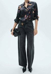 Mujer con una blusa negra de flores, vaqueros de pierna ancha de talle alto negros, botas de punta negra, sosteniendo un pequeño bolso de mano negro.
