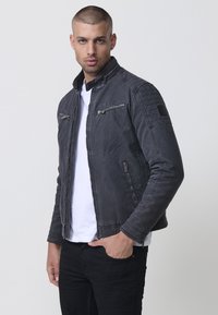 Fekete farmerdzseki álló gallérral, cipzáras zsebekkel, tűzött vállrészletekkel és karcsú szabással. Fehér póló fölött viselve.