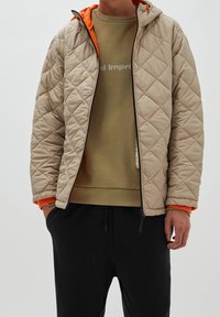 Veste matelassée beige avec capuche, doublure orange et fermeture éclair noire. Portée par-dessus un sweat-shirt vert et un pantalon noir.