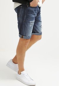 Man wearing rolled-up distressed denim shorts with hands in pockets and white sneakers standing against a plain background.