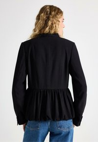 Sort peplumjakke lavet af glat stof, med en struktureret overdel, bløde samlede ved taljen og lange ærmer. Blå denim jeans synlige.