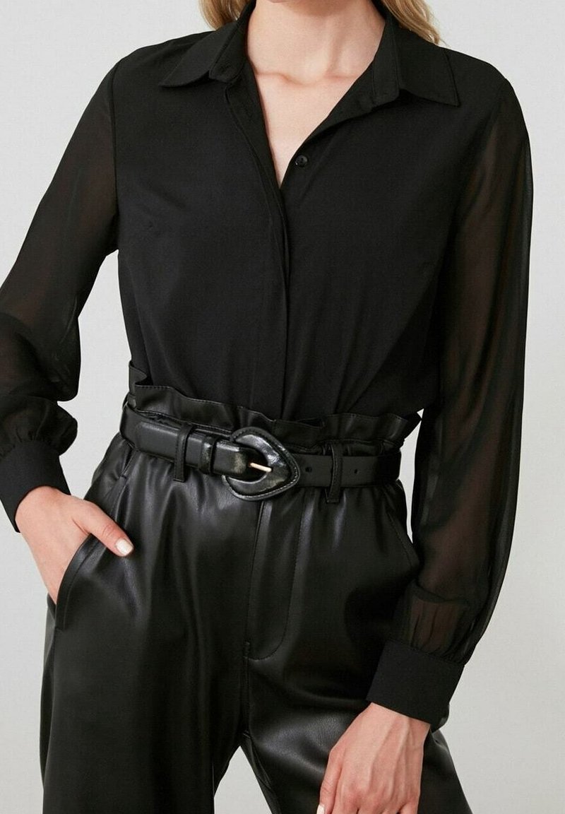 Blusa nera trasparente con maniche lunghe e colletto bottonato. Abbinata a pantaloni neri in ecopelle a vita alta e una cintura larga con una grande fibbia.