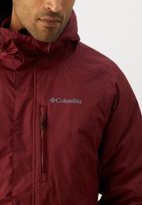 Maroon Columbia jacket with a hood, featuring a zippered chest pocket and the brand logo. Smooth texture with a lightweight design.