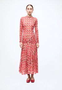 Robe à manches longues avec un motif abstrait rouge et crème, détail noué à la taille et jupe fluide ; tissu lisse et léger. Chaussures plates rouges portées.