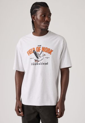 Mann mit geflochtenem Haar, der ein weißes T-Shirt mit orangefarbenem Text "FIELD OF WORK" und einer Illustration einer fliegenden Ente trägt.