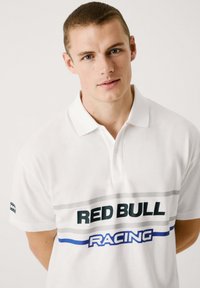 Balta polo marškinėliai su apykakle, „RED BULL“ ir „RACING“ logotipais žalios ir mėlynos spalvų, su pilkų dryžių juostele per krūtinę. Pagaminti iš medvilnės.