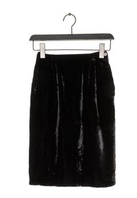 Černá sametová sukně visící na kovovém ramínku. Má hladkou texturu s jemným leskem, délku ke kolenům a minimalistický design.