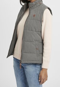 Graue gesteppte Weste mit hohem Kragen, Front-Reißverschluss und braunen Akzenten. Getragen über einem hellen Pullover mit abgerundetem Saum und blauen Jeans.