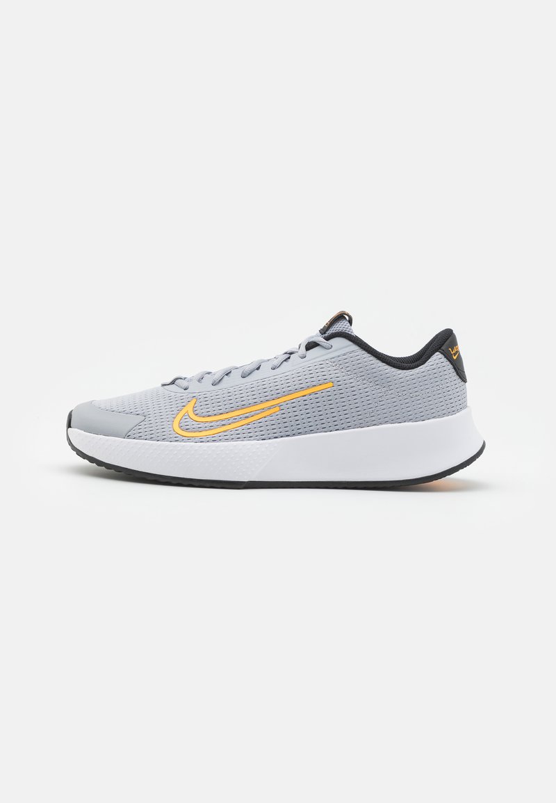 Scarpa sportiva grigia chiara con una tomaia in mesh texturizzato e suola in gomma bianca. Presenta un logo Nike giallo e dettagli neri sul tallone.