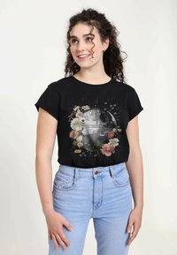 Sort T-shirt med et grafisk motiv af en Dødsstjerne omgivet af blomsterdesigns, med teksten "Star Wars" i gul nederst. Denim jeans er synlige.
