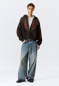 Veste à capuche noire zippée ornée de graphismes de flammes rouges, associée à un jean bleu délavé oversize et des chaussures noires épaisses. T-shirt gris en dessous.