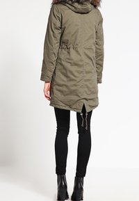 Olivgrön parkas med en konstläderhuva, med en insvängd midja, långa ärmar och ventilationsdetalj bak. Bärs över svarta leggings och ankelboots.