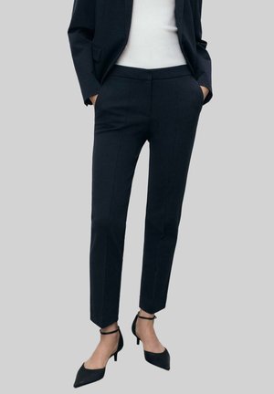 Kvinde iført sort slim-fit bukser, sort blazer, hvid top og sorte ankelstropper med hæl, stående med hænderne i lommerne.