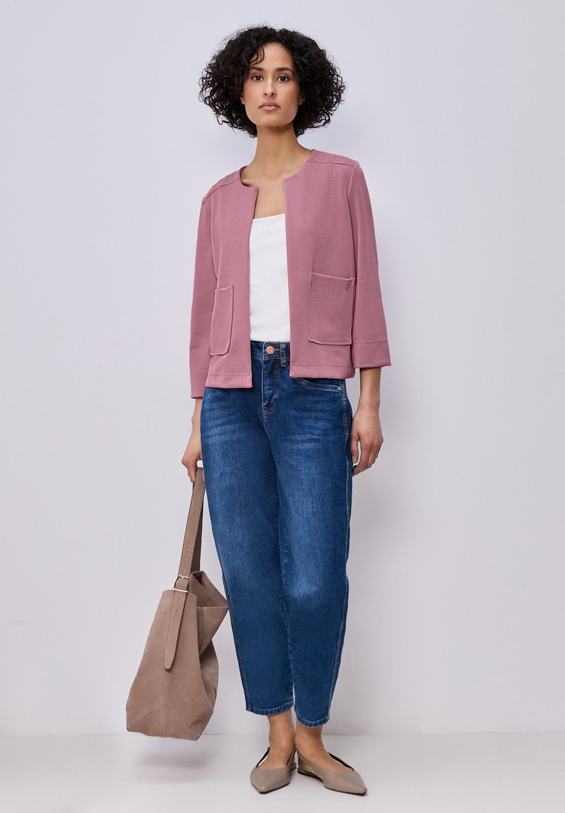 Mujer con cabello rizado, lleva una chaqueta malva, camiseta blanca, vaqueros azules, zapatos planos beige y sostiene un bolso tote grande beige.