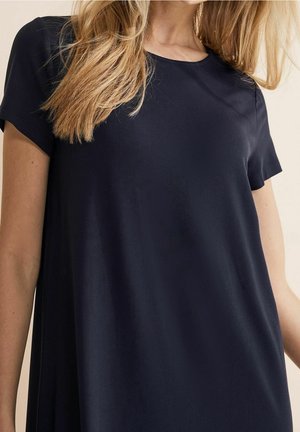 Kobieta ubrana w luźną sukienkę o krótkich rękawach w kolorze granatowym, z prostymi, blond włosami widocznymi na jasnym tle.