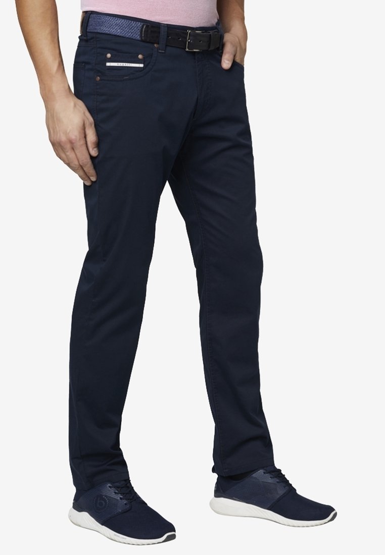 Muž v tmavě modrých kalhotách, odpovídajícím opasku a námořnických teniskách, stojí s jednou rukou v kapse a druhou u těla.