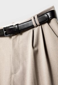 Pantalon beige à rayures avec un design plissé. Dispose d'une ceinture en cuir noir avec une boucle en métal argenté. Texture de tissu lisse.