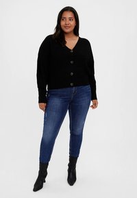 Cardigan nero a costine con scollo a V, caratterizzato da grandi bottoni marroni. Abbinato a jeans skinny di lavaggio medio e stivali neri alla caviglia.