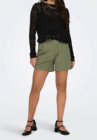 Femme portant un haut à manches longues en crochet noir, un short vert olive, des chaussures Mary Jane noires à talons, et un collier chaîne argenté, debout les mains détendues.