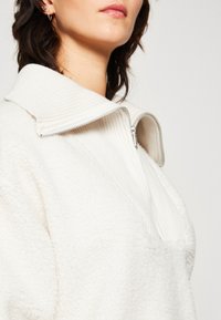Bílá fleecová mikina na zip s vroubkovaným límcem a texturovaným povrchem. Materiál je měkký, s nadměrným střihem a pohodlným designem.
