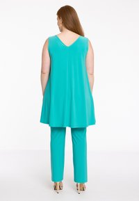 Een turquoise mouwloze tuniek met een V-rug, gecombineerd met bijpassende slim fit broek. De stof heeft een gladde textuur. Duidelijke details over fit en kleur.