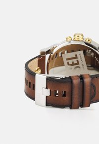 Orologio con cinturino in pelle marrone, caratterizzato da una cassa in acciaio inox argento, dettagli dorati, pelle texturizzata e una chiusura sicura in argento.