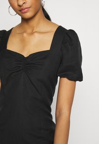 Abito nero con scollo quadrato, dettaglio arricciato sul petto e maniche corte a sbuffo. Tessuto morbido e testurizzato. Design semplice e aderente.