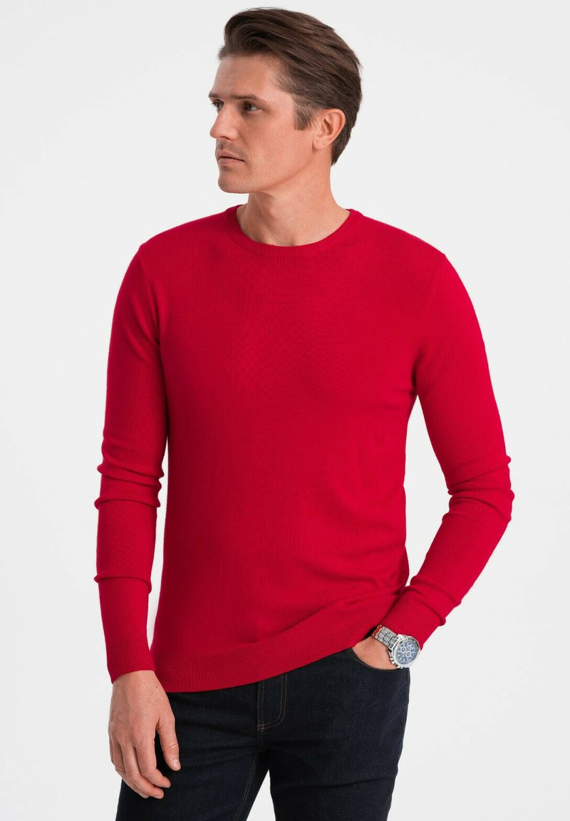 Pull long à manches longues en maille côtelée rouge avec un col rond, des manches ajustées et une texture lisse ; associé à un jean en denim foncé.