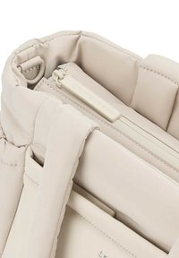Sac cabas matelassé beige avec fermeture éclair visible et tirette, avec des poignées cousues et un petit détail d’anneau interne.