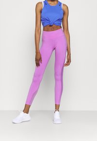 Top corto deportivo en azul con detalle de nudo, combinado con leggings rosa ajustados. La modelo lleva zapatillas deportivas blancas. Tejido suave y elástico.