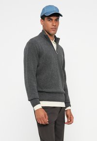 Grauer Strickpullover mit einem Viertelreißverschluss, gerippten Bündchen und Saum. Über einem weißen Hemd getragen, kombiniert mit einer hellblauen Mütze.
