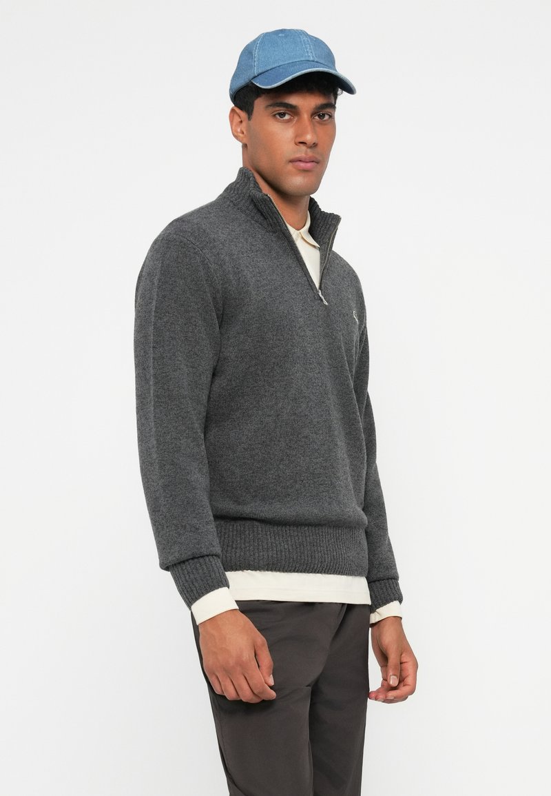 Grå stickad tröja med kvartdragkedja, ribbade ärmslut och nederkant. Buren över en vit skjorta, kombinerad med en ljusblå keps.