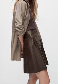 Gonna in pelle marrone con un design svasato, abbinata a un blazer in tessuto liscio color taupe e a un top aderente viola, che mette in mostra un look a strati.