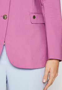 Różowy żakiet o dopasowanym kroju, zapinany na pojedynczy guzik, z kieszenią ozdobioną metalowym akcentem. Gładka faktura i czyste linie.