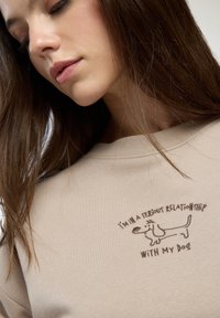 Beżowa bluza z wyszywaną ilustracją psa i napisem: "Jestem w poważnym związku z moim psem." Miękka tekstura.