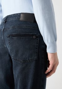 Acercamiento de una persona que lleva pantalones vaqueros de un azul oscuro con una etiqueta de cuero negro de la marca y un logotipo metálico en el bolsillo trasero, combinados con una camiseta de manga larga de un azul claro.
