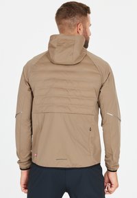 Leichte, braune Jacke mit Kapuze, mit einem gesteppten Rücken, reflektierenden Akzenten und elastischen Bündchen. Enthält eine mit Reißverschluss versehene Tasche an der Seite.