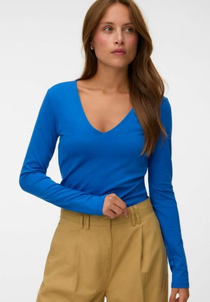 Femme aux longs cheveux bruns portant un haut bleu vif à col en V manches longues et un pantalon beige taille haute sur un fond uni.