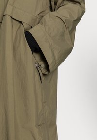 Abrigo largo color verde oliva hecho de tela ligera y arrugada, con un bolsillo lateral con cremallera y mangas anchas y sueltas.