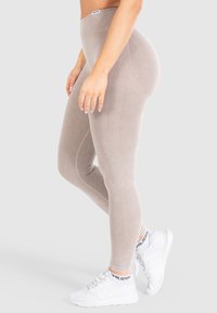 Beżowe legginsy z wysokim stanem o prążkowanej teksturze, z pionowymi paskami po bokach, w połączeniu z białymi butami sportowymi.