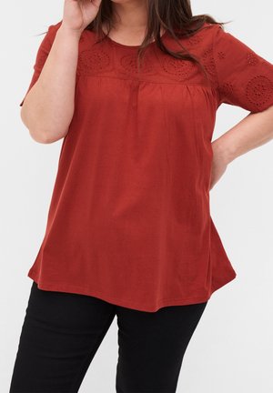 Femme portant un chemisier rouge ample avec des motifs circulaires brodés sur la poitrine et les manches, associé à un pantalon noir.