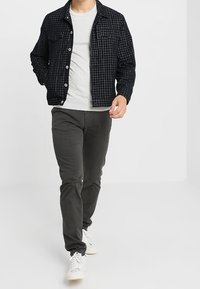 Dunkelkarierte Jacke mit Knopfverschluss und zwei Brusttaschen über einem hellgrauen Hemd, kombiniert mit dunklen Hosen und weißen Turnschuhen.