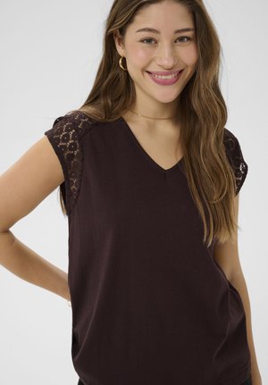 Young woman smiling, wearing a dark brown V-neck top with lace cap sleeves and gold hoop earrings.